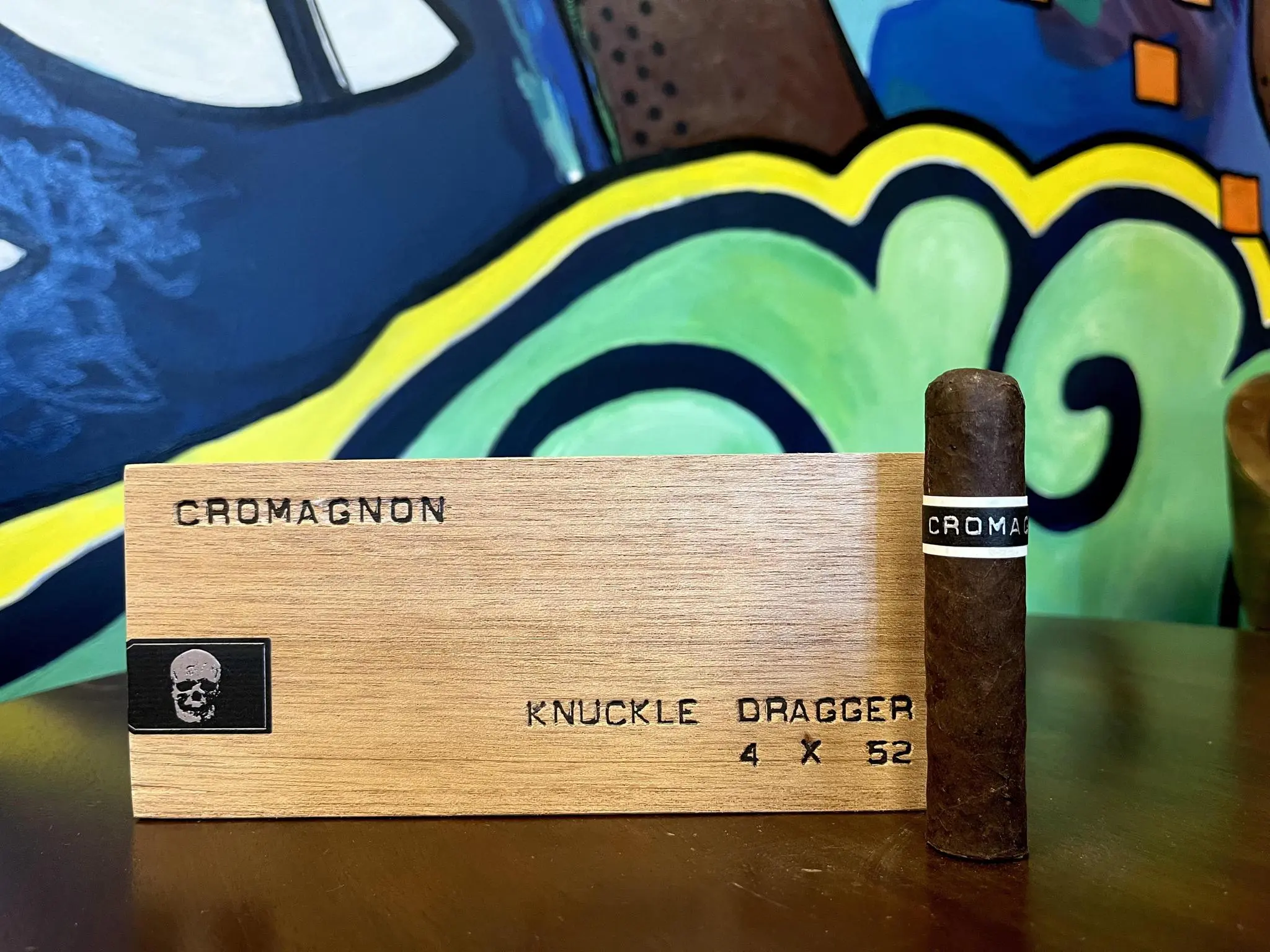 RoMa Craft CroMagnon Knuckle Dragger cigar displayed next to its wooden box featuring a skull logo and 'KNUCKLE DRAGGER 4 X 52' branding against colorful graffiti background