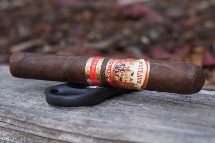 A dark, oily AJ Fernandez Enclave Broadleaf Toro cigar resting on weathered wooden boards, displaying its rich Connecticut Broadleaf wrapper and distinctive red and gold band.