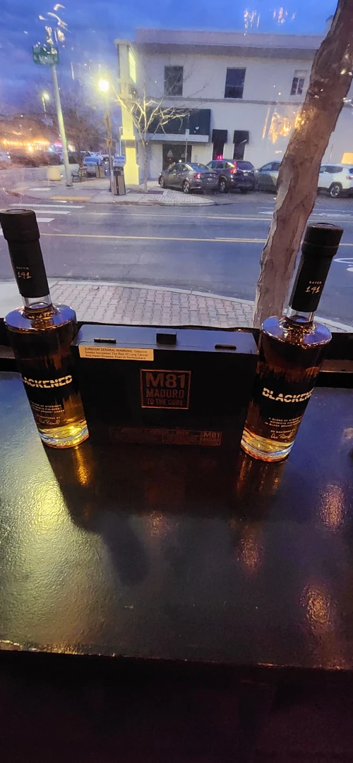 Two bottles of Blackened American Whiskey flanking an M81 Maduro to the Core cigar box on a table by a window at dusk.