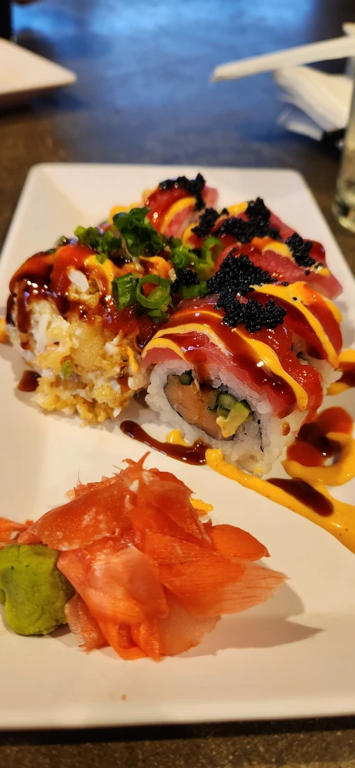 A plated specialty sushi roll topped with tuna, eel sauce, spicy mayo, green onions, and tobiko, served with pickled ginger and wasabi.