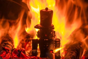 Intense orange and yellow flames engulfing metal objects in a blacksmith's forge, with glowing hot coals visible at the base