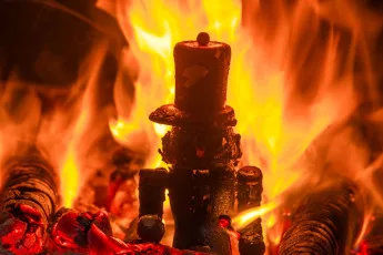 Intense orange and yellow flames engulfing metal objects in a blacksmith's forge, with glowing hot coals visible at the base