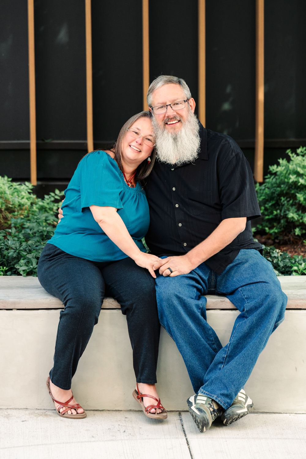 William and Carolyn Brown of Brown's Life sitting close together on a bench, smiling, holding hands in front of a dark wall with green plants.